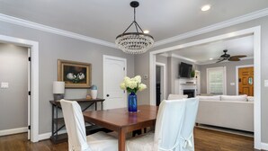 A dining area with a wooden table, white chairs, a chandelier, a vase with flowers, and a painting on the wall. - View Street Retreat | Pet Friendly, Fenced Yard, Walk to Town! (Black Mountain)