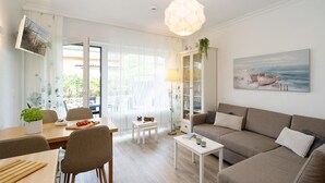 A modern living room with a sofa, dining table, and chairs. There is a bookshelf, a lamp, and a painting on the wall. - Syltoase - 3-zimmer App. für 4 Personen mit Terrasse, Stellplatz, Wlan. Haustier Erlaubt (Westerland)