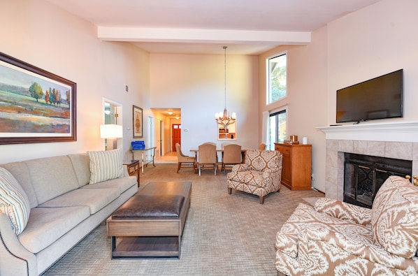 A living room with a sofa, ottoman, patterned armchair, and a fireplace. There is a television mounted above the fireplace and a painting on the wall. - Private Golf Resort Setting 3 bedrooms (Napa)