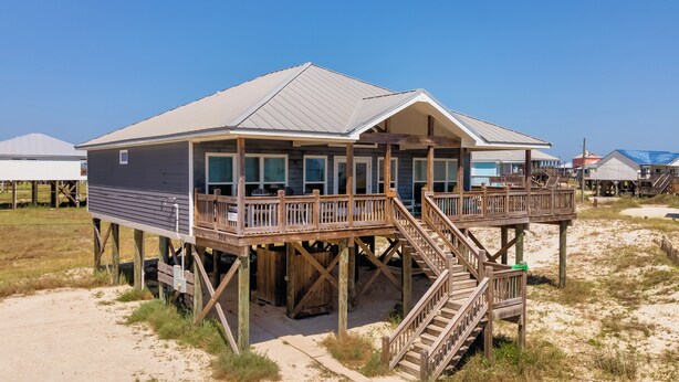 A wooden house with a balcony and stairs on stilts, surrounded by sand and grass. - Tidewater East | Gulf-Side Comfort & Style (Dauphin Island)