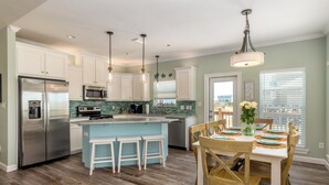 A modern kitchen with white cabinets, stainless steel appliances, a blue island with stools, and a dining area with wooden chairs and a table set for four. - Tidewater East | Gulf-Side Comfort & Style (Dauphin Island)