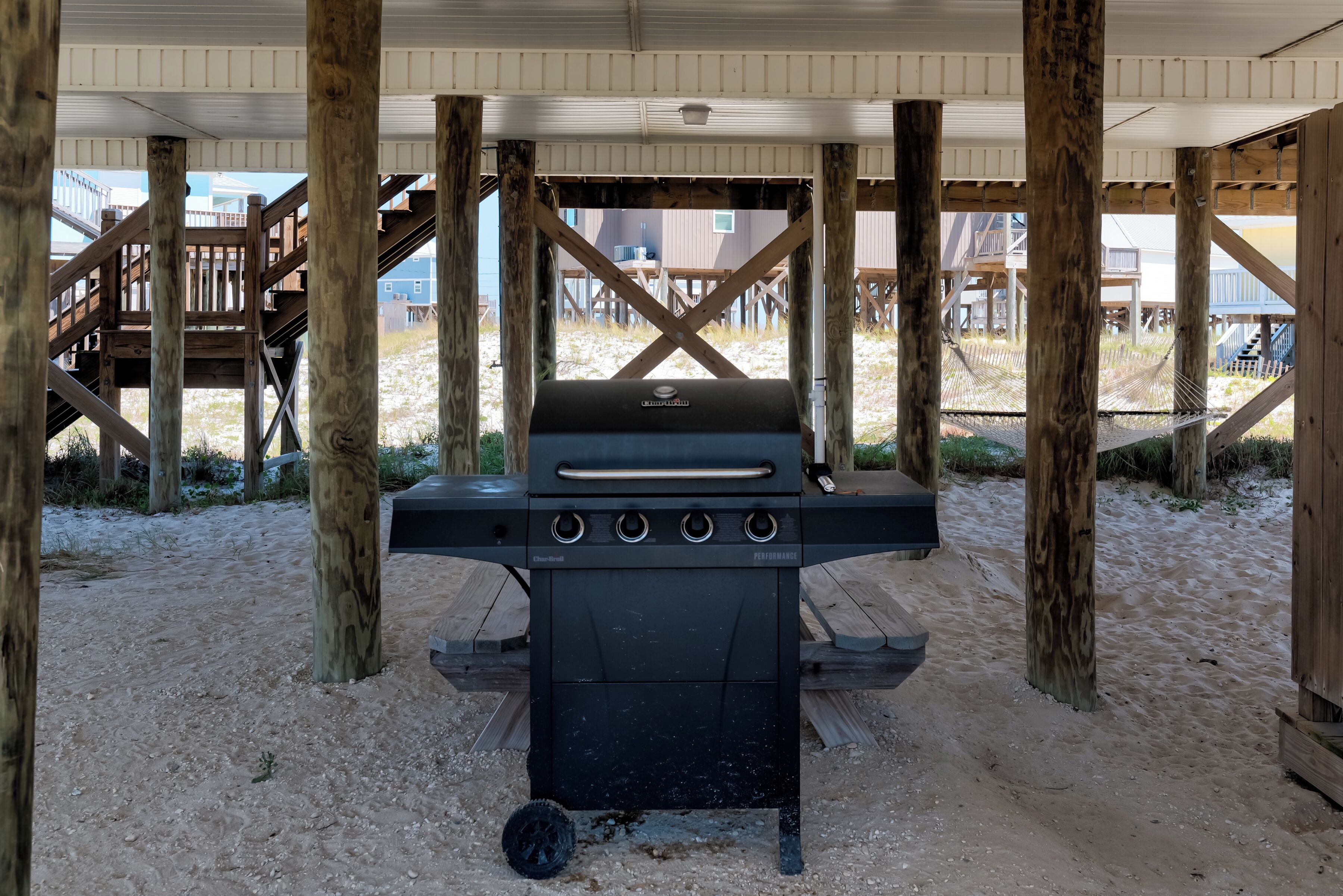 Una parrilla para barbacoa debajo de una terraza de madera, sobre un suelo arenoso.