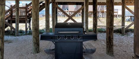 Una parrilla para barbacoa debajo de una terraza de madera, sobre un suelo arenoso.