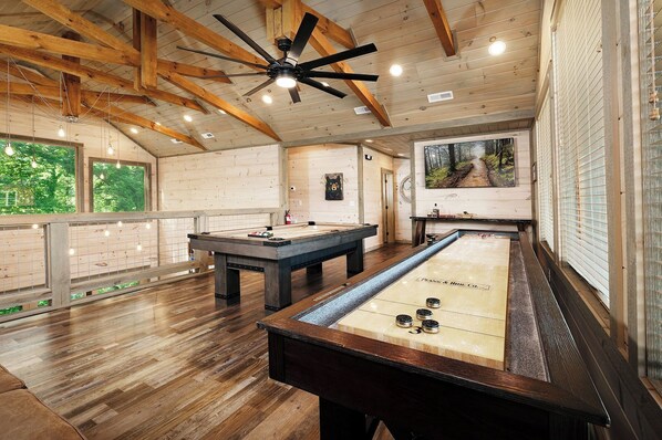 A spacious indoor pool table area with wooden flooring and ceiling beams, a ceiling fan, and a large window offering a view of greenery.