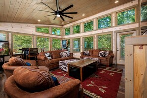 A spacious living room with a high ceiling, large windows, and a ceiling fan.