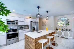 A modern kitchen with stainless steel appliances, a wooden island with bar stools, pendant lighting, and decorative wall art. - Tropical Miami/Heated pool/Basketball Court/Pin Pong Table/Patio Chess/Mini Golf (Miami)
