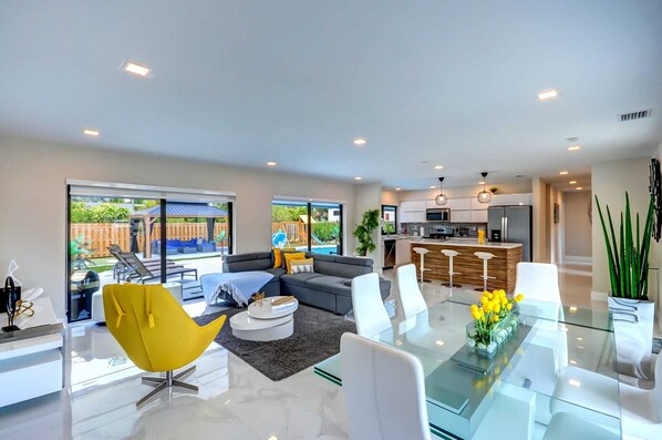 A modern living room with a glass dining table, a sofa, and a yellow chair. There is a kitchen area with a bar and a large window. - Tropical Miami/Heated pool/Basketball Court/Pin Pong Table/Patio Chess/Mini Golf (Miami)