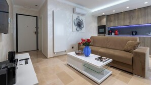 Una sala de estar moderna con un sofá color beige, una mesa de centro con un jarrón de flores rojas, un televisor de pantalla plana y una zona de cocina con armarios de madera e iluminación LED azul.
