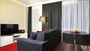 A modern hotel room with a flat-screen TV, a sofa, a small round table with fruit, and a lamp.
