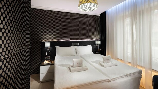 A hotel room with a large bed, two bedside tables, a headboard with a geometric pattern, and a chandelier.