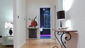 Un pasillo con suelo de madera, un reloj negro en la pared, una mesa con lámpara y un cuadro de una rosa roja.