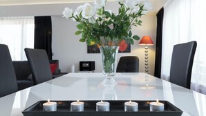 A modern hotel room with a glass-top dining table, a vase of white flowers, and a tray of lit candles.