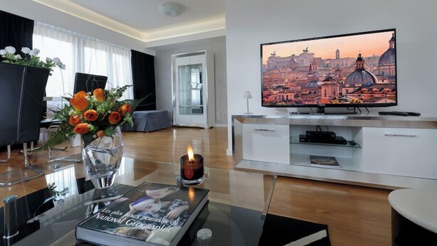 A modern living room with a large flat-screen TV displaying a cityscape, a glass coffee table with a book, and a vase with flowers.