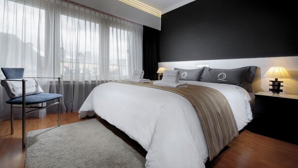 Una camera d'hotel moderna con un letto grande, una sedia, un comodino con una lampada e tende leggere alla finestra.