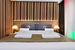 Quarto de hotel com uma cama grande, cortinas listradas, duas luminárias de cabeceira e toalhas com a marca "The Queen Luxury Living".