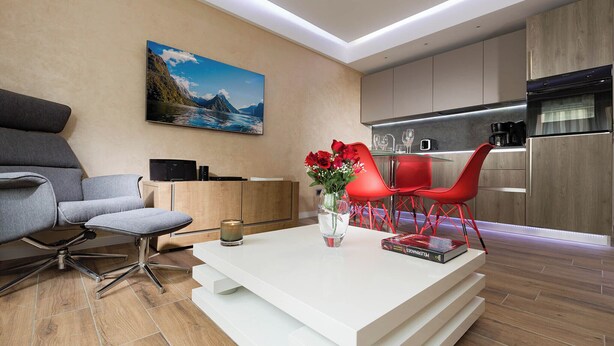 A modern living room with a white coffee table, grey sofa, red chairs, and a painting of a coastal landscape.