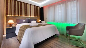 A modern hotel room with a large bed, bedside tables, a chair, and a window with sheer curtains.