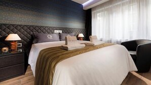 A hotel room with a large bed, two bedside tables with lamps, a dark headboard, and a window with sheer curtains.