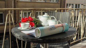 Ein runder Wickertisch mit weißem Teekessel, Tasse, Untertasse und einer roten Rose auf einem Balkon.