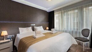 Habitación de hotel con una cama grande, una mesita de noche con lámpara, una silla y una ventana con cortinas traslúcidas.