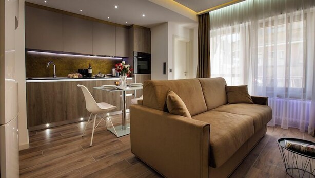 A modern living room with a sofa, a dining table, and a kitchen area with wooden cabinets and a built-in oven. - Carlotta -33 Appartement avec Balcon (Luxembourg)