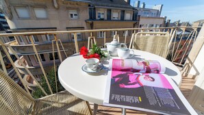 Una mesa redonda con una revista, una tetera, tazas y una flor roja en un balcón con vistas a la ciudad.