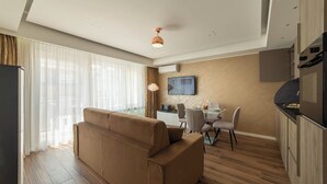 Un salon moderne avec un canapé, un coin repas comprenant une table et des chaises, une cuisine équipée d’un réfrigérateur et d’un four, et un téléviseur fixé au mur.