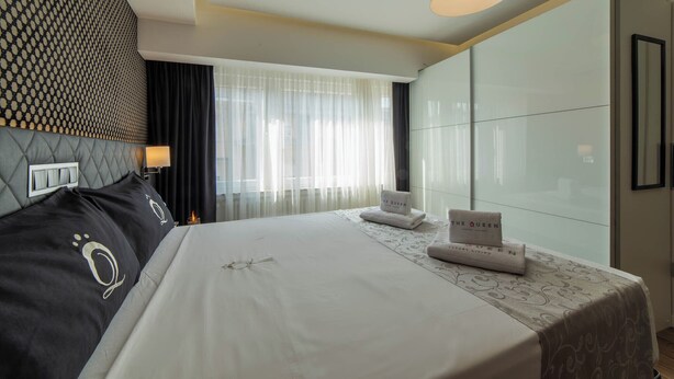 Une chambre d’hôtel avec un grand lit, une tête de lit ornée d’un motif géométrique, une table de chevet avec une lampe et une fenêtre avec des rideaux légers.