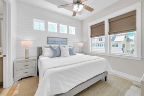 A bedroom with a bed, nightstands, a ceiling fan, a large window with blinds, and a view of a house.