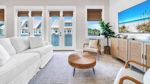 A modern living room with a white sofa, a round wooden coffee table, a wicker cabinet, and a large TV screen displaying a beach scene.