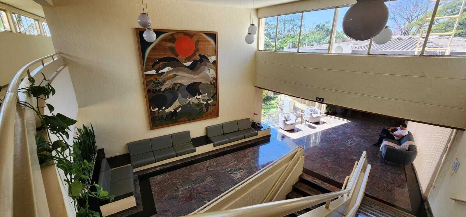 Um hall de hotel moderno com uma receção de mármore, uma área de estar com sofás, uma obra de arte de grandes dimensões retratando uma cena tradicional, e vista para o exterior através de grandes janelas.
