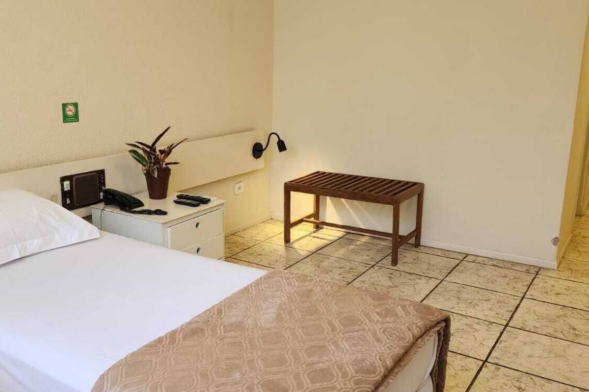 Ein Hotelzimmer mit einem Einzelbett, einem Nachttisch mit Telefon und Pflanze, einem kleinen hölzernen Hocker und einer Wandlampe.