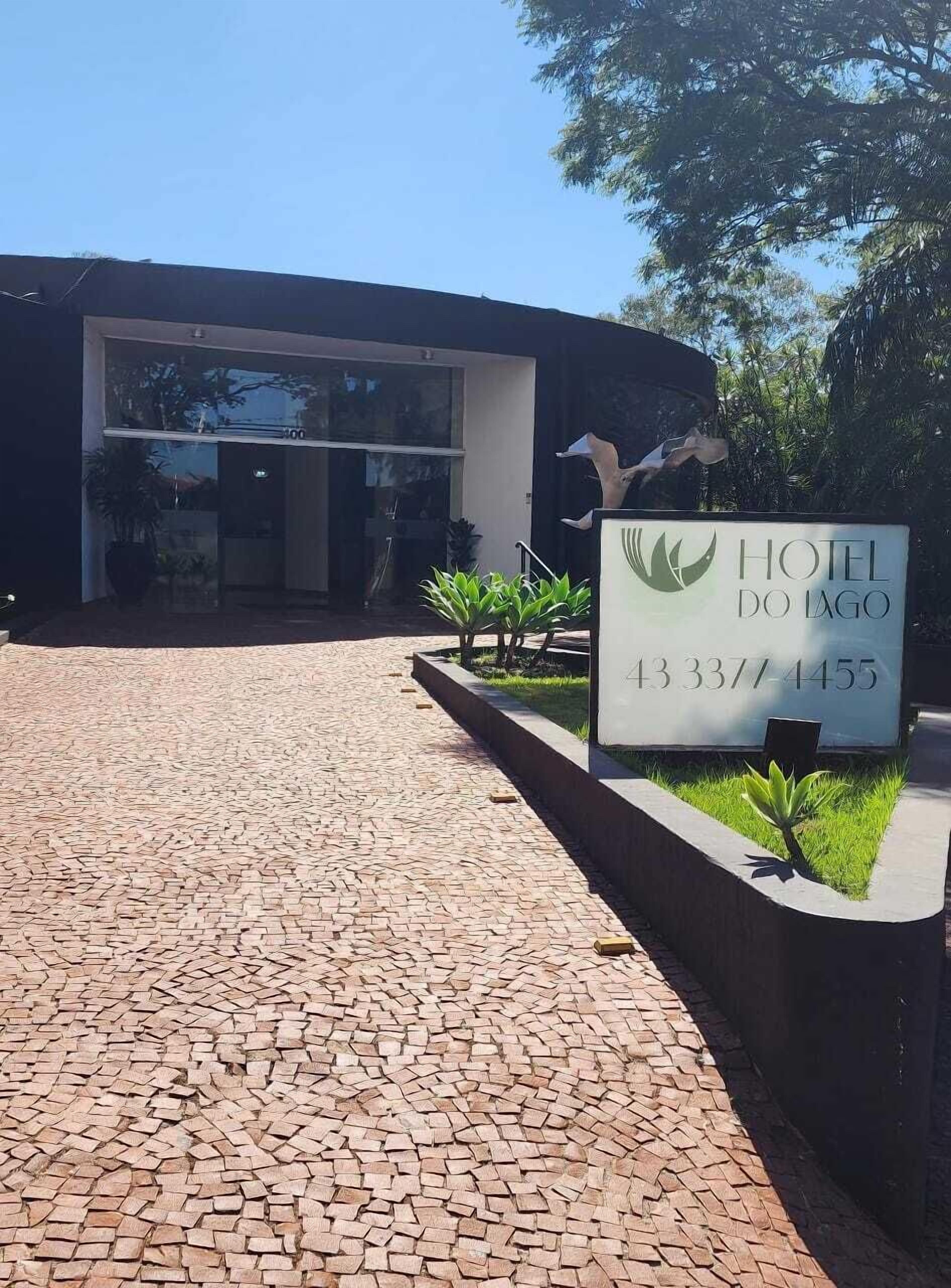 Una entrada de hotel con un camino pavimentado que conduce a un edificio moderno. Un cartel muestra el nombre del hotel y su número de contacto.
