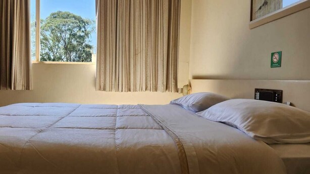 A bedroom with a large bed, beige curtains, a window with a view of trees, and a small table. - Goldmen Business Londrina (Londrina)