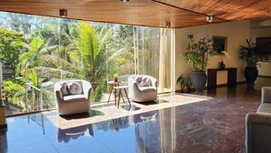 Una habitación amplia con un gran ventanal que da a un jardín tropical, suelo de mármol y una zona de estar con dos sillones y un sofá.