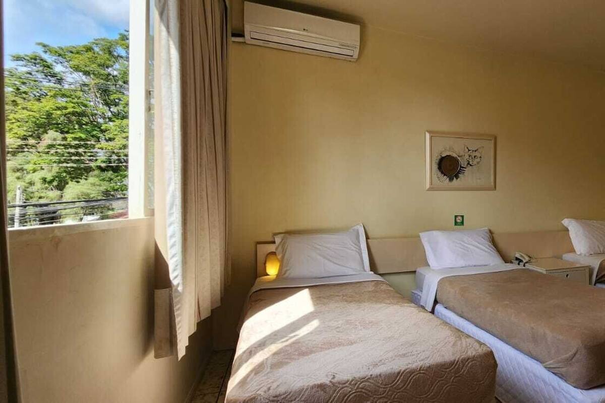 Ein Hotelzimmer mit zwei Einzelbetten, einem Nachttisch mit Lampe, einer Wand-Klimaanlage und einem Fenster mit Vorhängen.