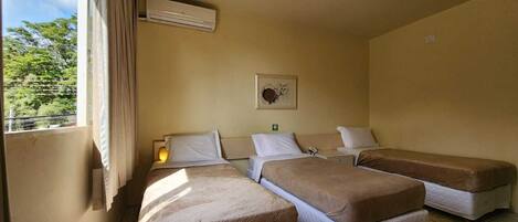 A hotel room with two single beds, a bedside table with a lamp, a wall-mounted air conditioner, and a window with curtains.