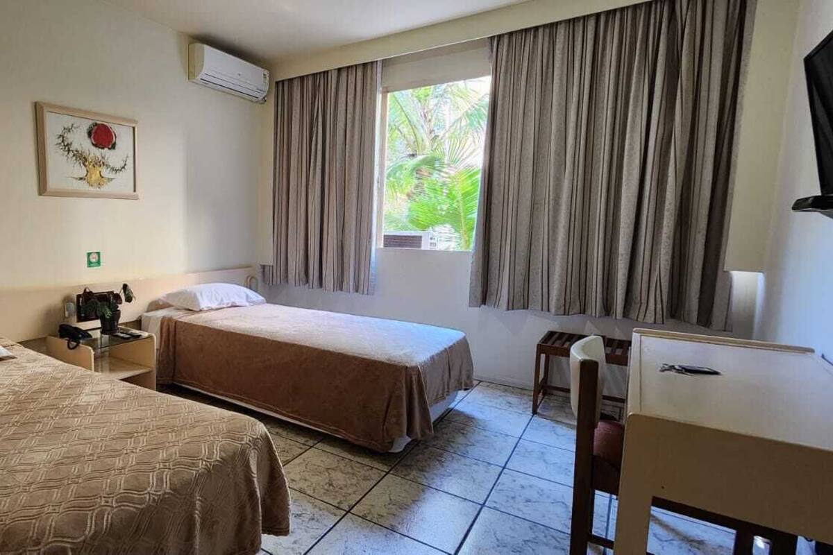 Ein Hotelzimmer mit zwei Betten, einem Schreibtisch, einem Stuhl, einem Fenster mit Vorhängen und einer Wandklimaanlage.