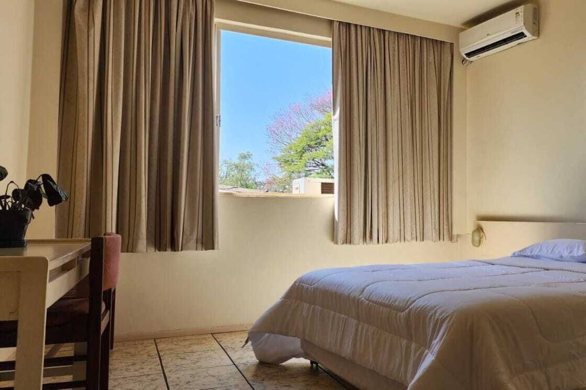 Ein Hotelzimmer mit Bett, Schreibtisch, Stuhl, einem Fenster mit Vorhängen, einer Klimaanlage und einem an der Wand montierten Telefon.