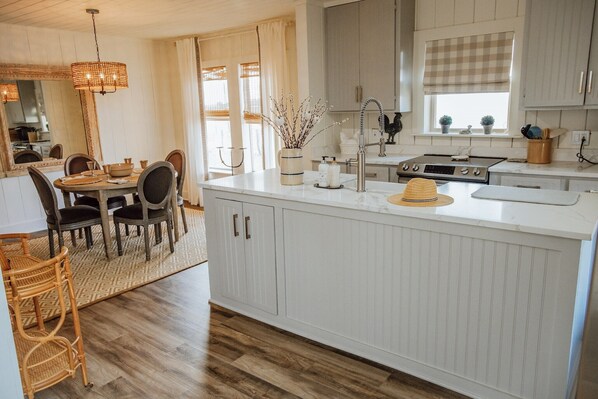 A modern kitchen with white cabinetry, a central island, and a dining area with a wooden table and chairs. - MoonShiner Farm - Shiner TX (Shiner)