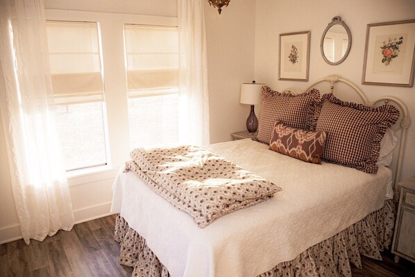 A neatly made bed with a floral-patterned blanket and pillows, a bedside table with a lamp, and a window with sheer curtains. - MoonShiner Farm - Shiner TX (Shiner)