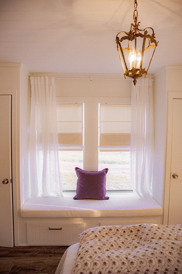 A bedroom with a window seat, a bed with a patterned bedspread, and a chandelier. - MoonShiner Farm - Shiner TX (Shiner)