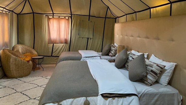 A tent-style room with two beds, a small table, and a wicker chair. - Arena Desert Camp & Activitys (Merzouga)