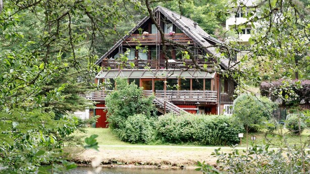 A rustic cabin with a wooden exterior, a balcony, and a pond in the foreground. - Holiday flat "am Talsee", with lake view, balcony and Wifi (Unterkirnach)