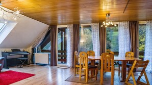 A dining area with a wooden table and chairs, a piano, and large windows with curtains. - Holiday flat "am Talsee", with lake view, balcony and Wifi (Unterkirnach)