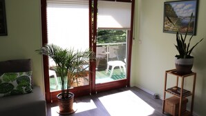 Une pièce dotée d’une porte-fenêtre coulissante donnant sur un balcon, d’une plante en pot, d’une petite étagère en bois et d’un tableau encadré accroché au mur.