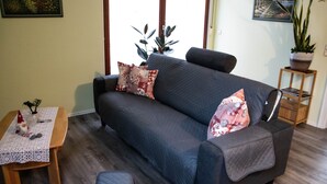 Un salon avec un canapé de couleur foncée, des coussins à motifs, une petite table en bois recouverte d’une nappe blanche et une plante en pot.