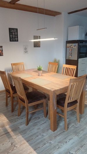 Une table à manger en bois, accompagnée de six chaises, un luminaire suspendu, un réfrigérateur et un mur sur lequel est affiché un menu de cafés.