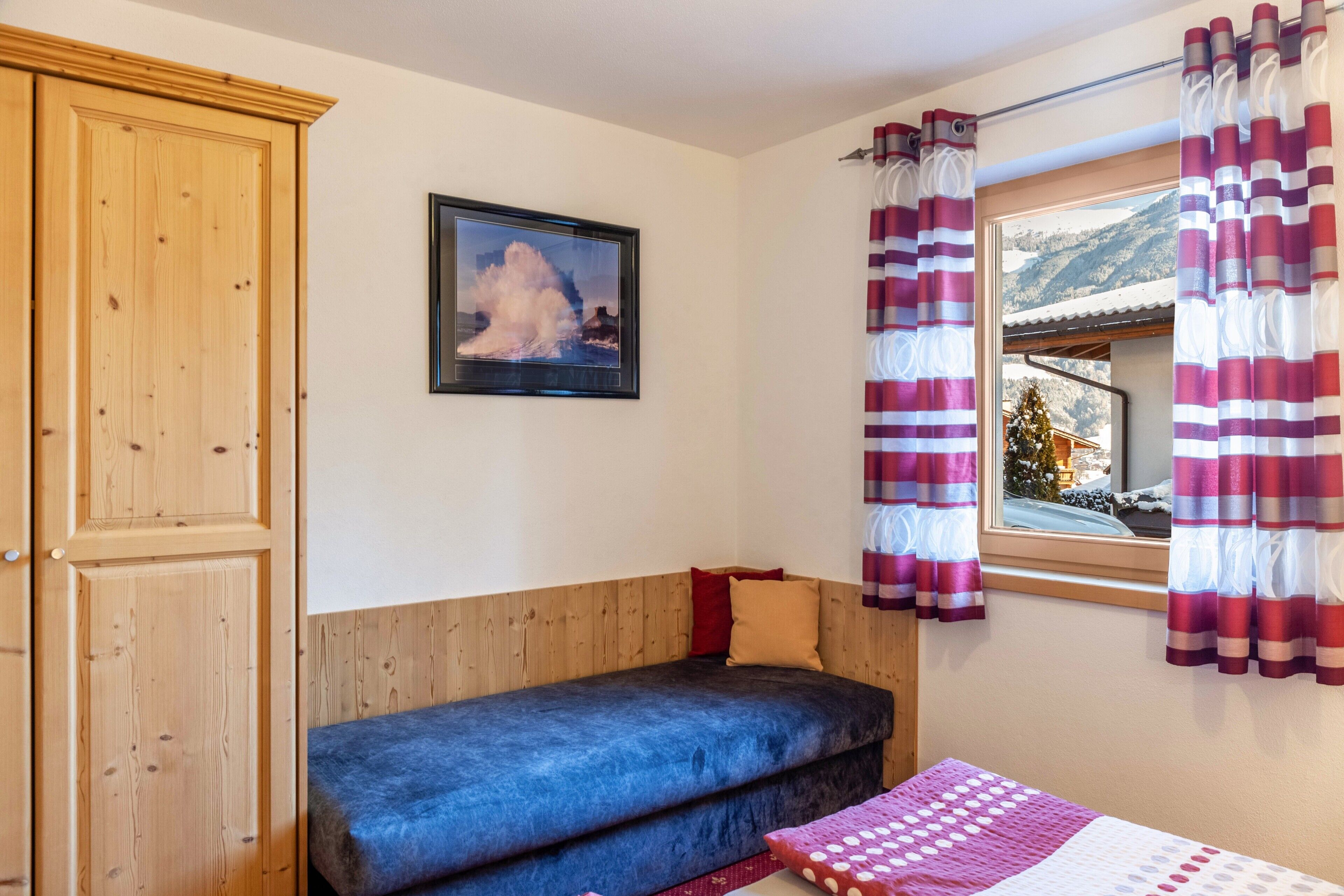 A room with a wooden wardrobe, a bed with a blue mattress, a window with curtains, and a framed picture on the wall.