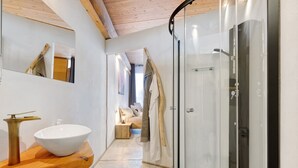 Un baño moderno con tocador de madera, un lavabo blanco grande, cabina de ducha de cristal y una habitación visible al fondo.